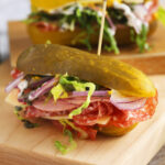 A fully assembled pickle sandwich with salami, cheese, lettuce, onion, and tomato, all sandwiched between the halves of a large pickle and held together with a sandwich pick.