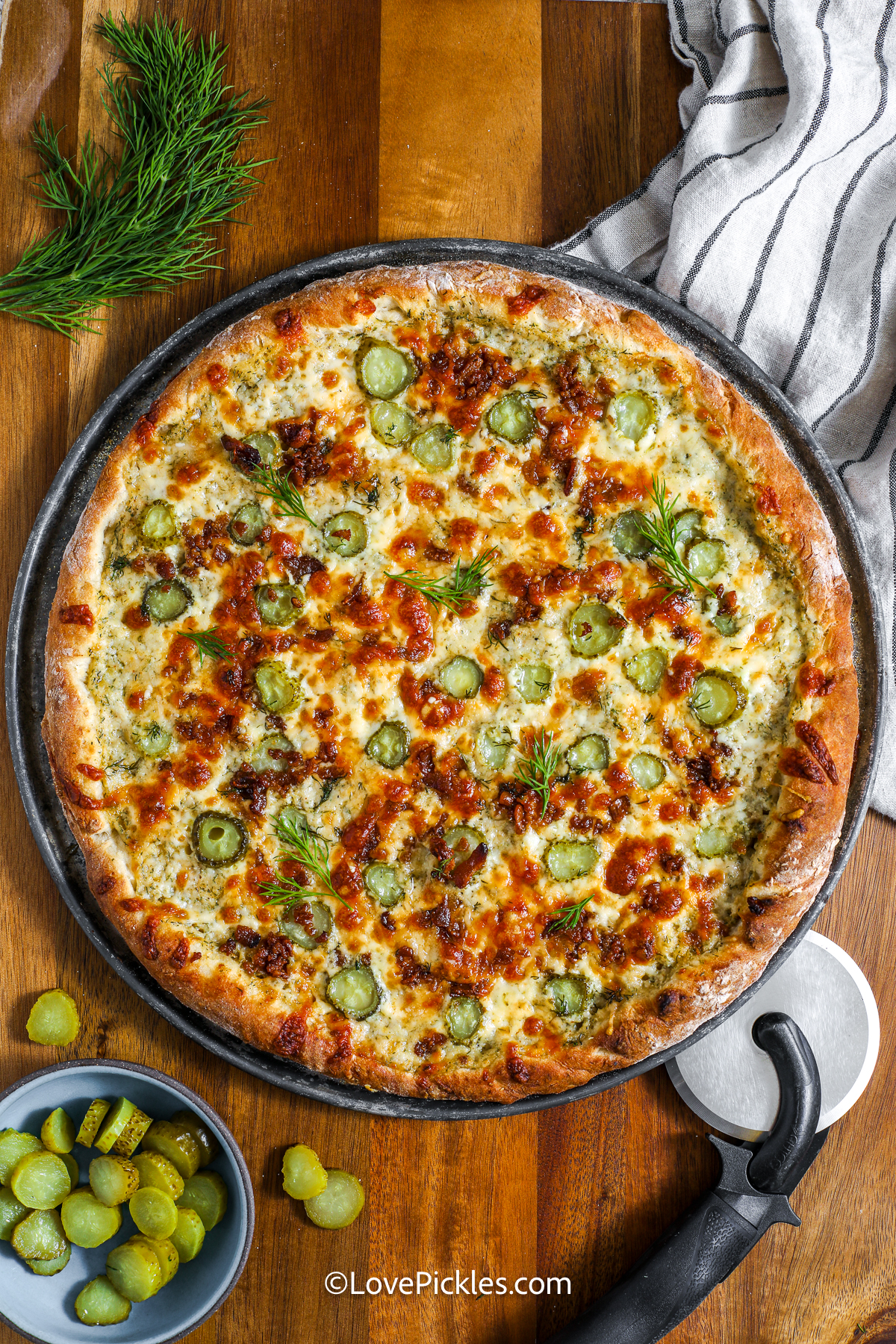Dill pickle pizza brings everything we love: cheese, pickles, and creamy goodness all together in one slice! A homemade alfredo sauce is seasoned just right and spread over pizza dough, then topped with cheese, bacon, and pickles galore. After baking, a sprinkle of dill gives it a fresh pop. Whether you're team pickle or just love fun flavors, this pizza is for you! #lovepickles cooked dill pickle pizza