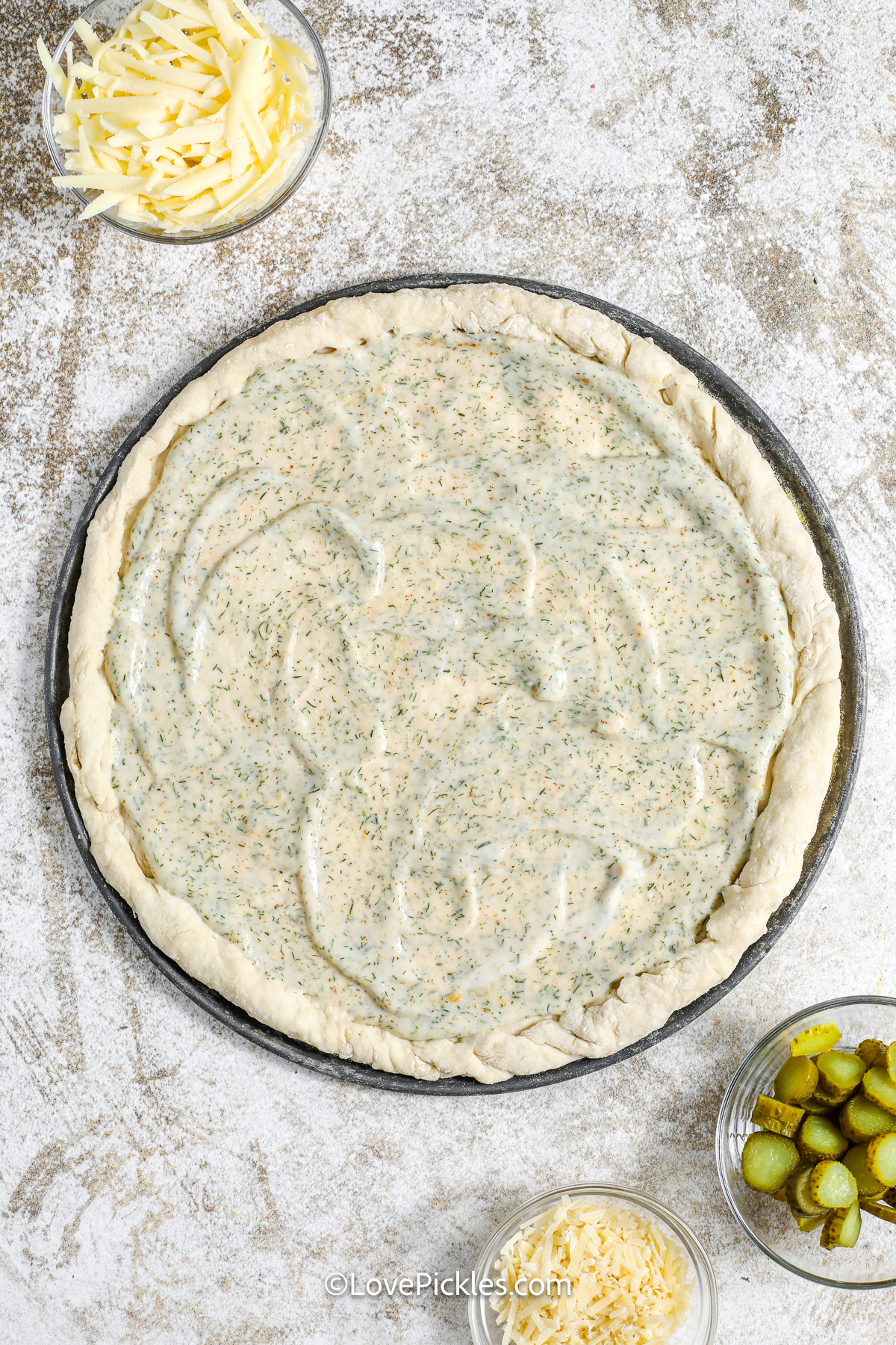 This dill pickle pizza is a tangy twist on white pizza. The Alfredo sauce is creamy with just the right kick of dill and spice, and it's topped with melted cheese, smoky bacon, and tart pickle slices. Bake it up crispy and finish with fresh dill for fun on pizza night. It’s a bold and delicious way to shake things up in the kitchen! #lovepickles sauce spread on pizza dough to make dill pickle pizza.