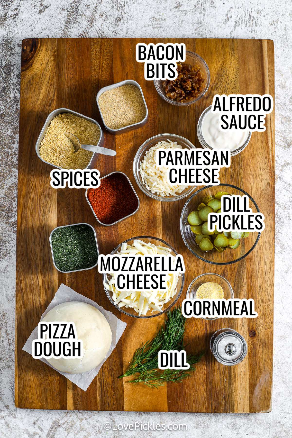 If you love pickles, you need to try dill pickle pizza! Creamy alfredo sauce gets jazzed up with dill and spices, then it's layered with gooey cheese, crispy bacon, and a generous handful of sliced pickles. Baked until the crust is golden and crisp, it’s finished with fresh dill for a bright, herbal kick. Every slice is packed with bold, savory flavor you’ll crave repeatedly. #lovepickles ingredients assembled to make dill pickle pizza, including spices, bacon bits, parmesan cheese, mozzarella cheese, dill pickles, cornmeal, alfredo sauce, and pizza dough