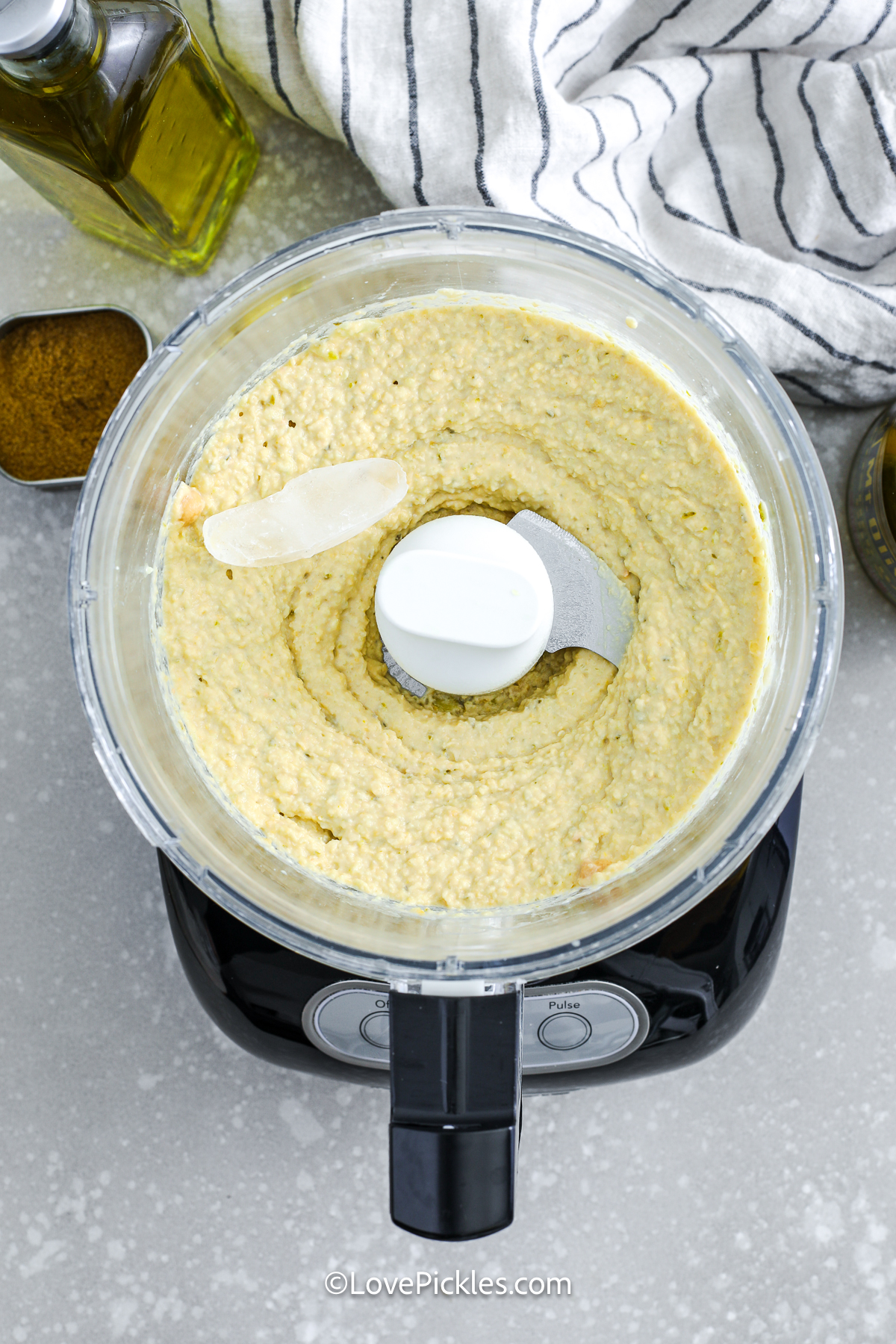 Adding an ice cube to blended dill pickle hummus in a food processor