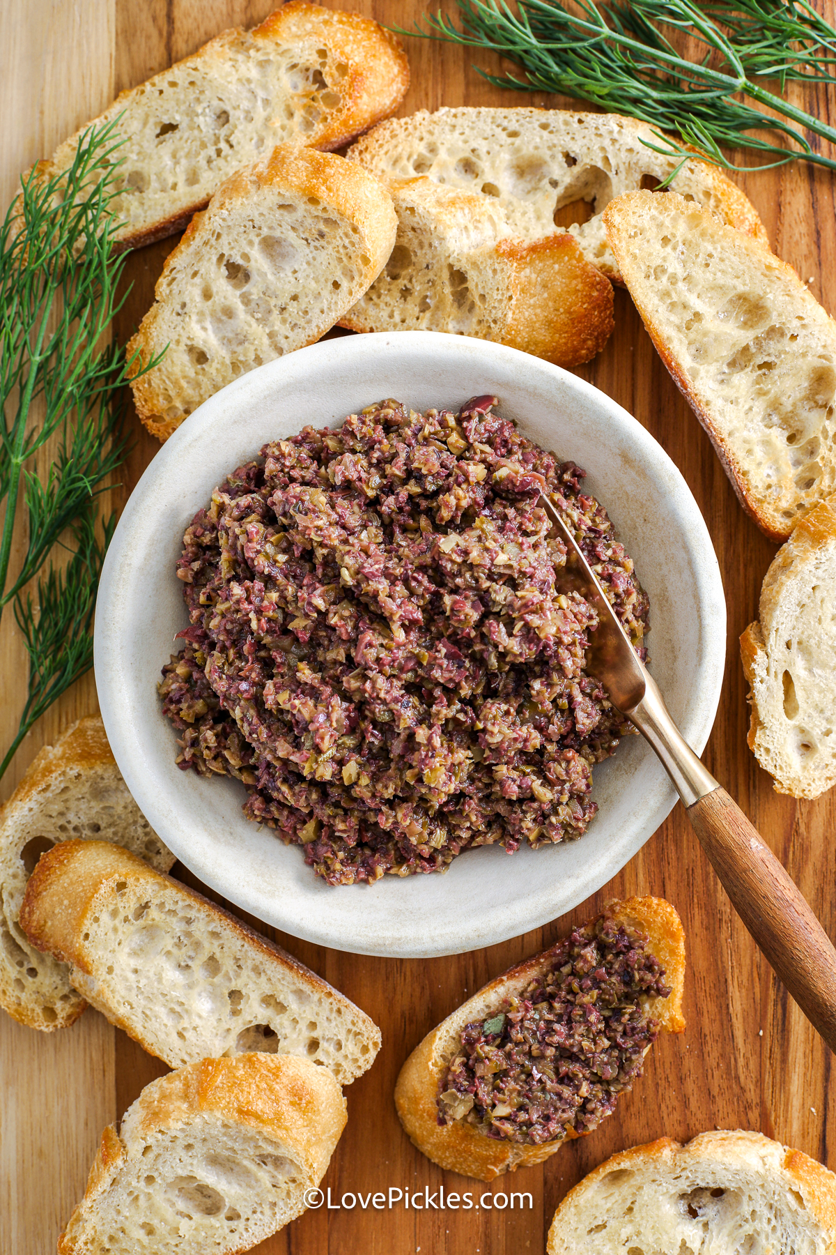 This dill pickle tapenade brings together salty olives, zesty pickles, and briny capers into a flavorful, textured spread. Garlic and anchovy add savory depth, while lemon juice adds brightness to balance the richness. Olive oil binds it all into a luscious paste perfect for spreading on crusty French bread or crunchy crostini. Great as an appetizer or sandwich topping, it’s a unique way to enjoy pickles beyond the jar. #lovepickles dill pickle tapenade recipe in a bowl with a knife