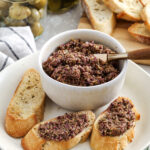 dill pickle tapenade recipe on bread slices with olives, bread, and pickles in background