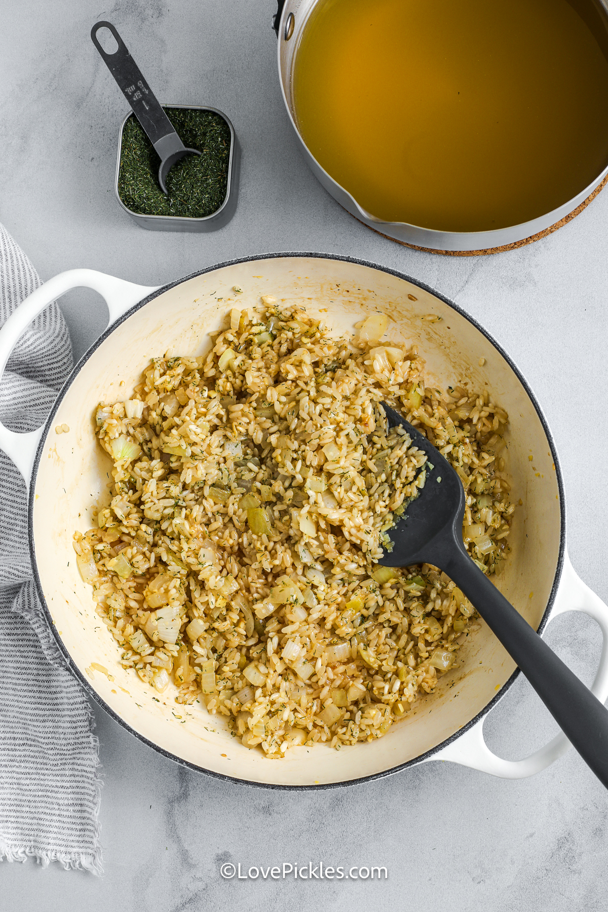 Dill risotto is rich, creamy, and full of zippy dill flavor that shines in every bite. Sautéed onions and garlic start the base, while the rice simmers in warm broth and tangy pickle juice. A touch of wine adds complexity, and butter and cheese give it a smooth, luscious finish. This risotto is bright, savory, and endlessly comforting making it perfect for pickle fans and flavor seekers alike. #lovepickles cooking dill risotto in a pot